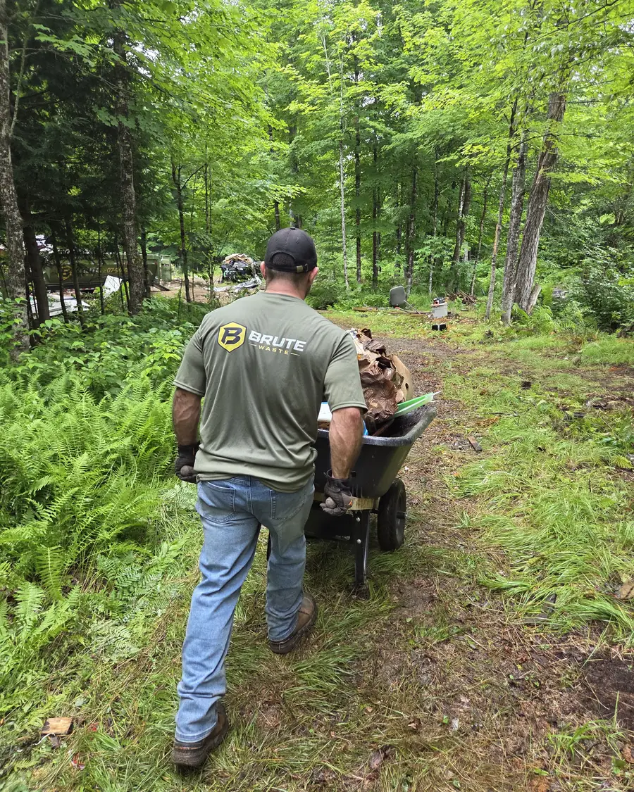 A Brute Waste crew member pushing a loaded wheelbarrow along a wooded path