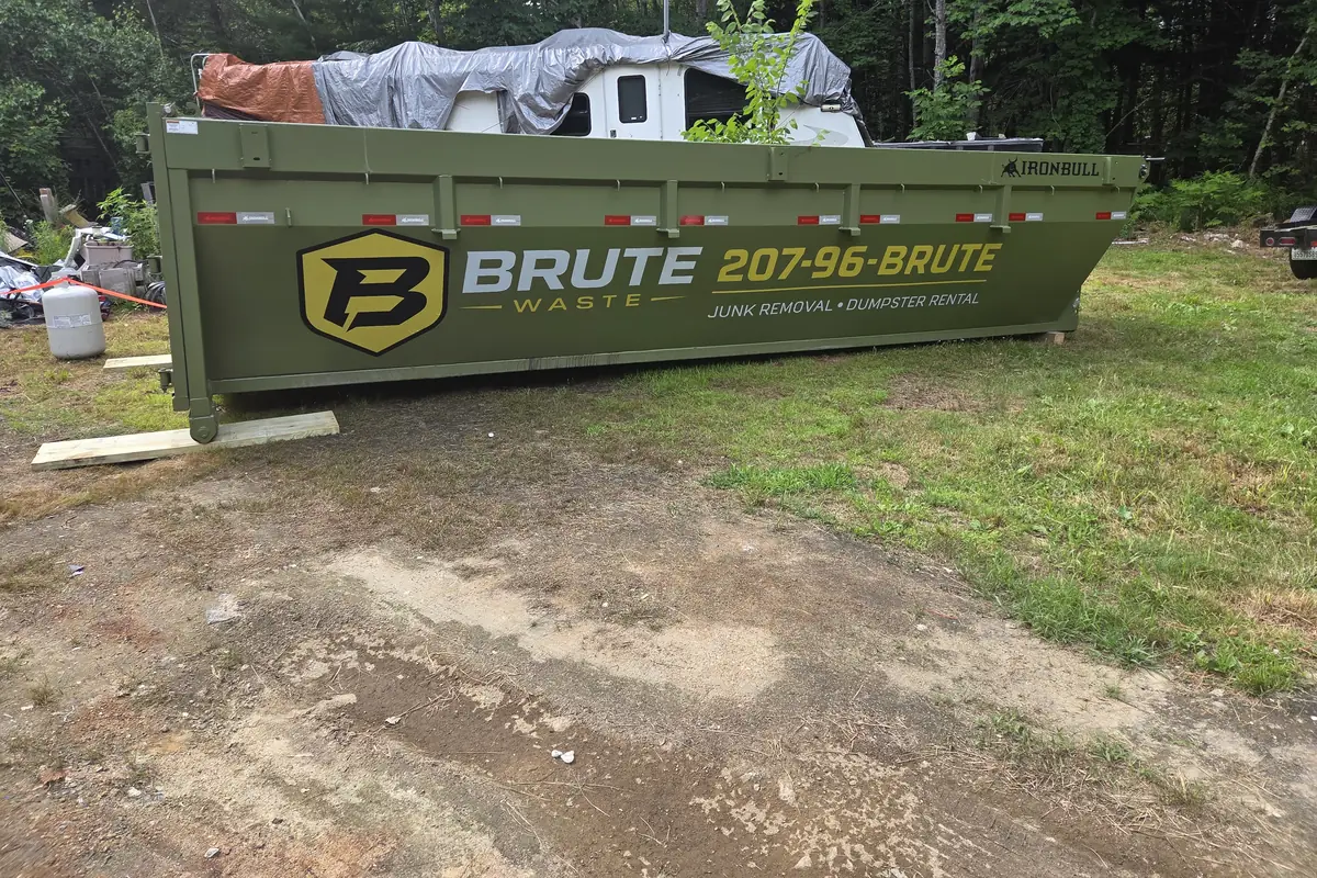 A Brute Waste roll-off dumpster parked on bare ground beside a covered camper