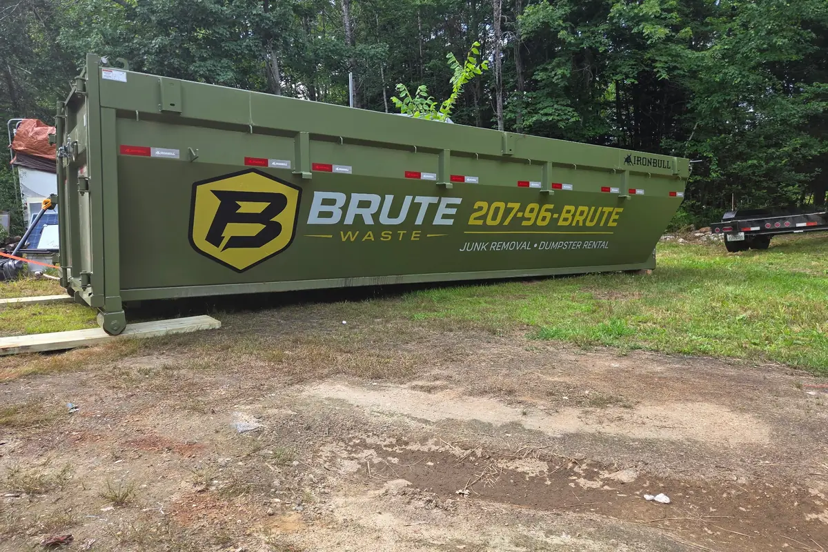 A green Brute Waste roll-off dumpster standing in a cleared patch of Maine woodland