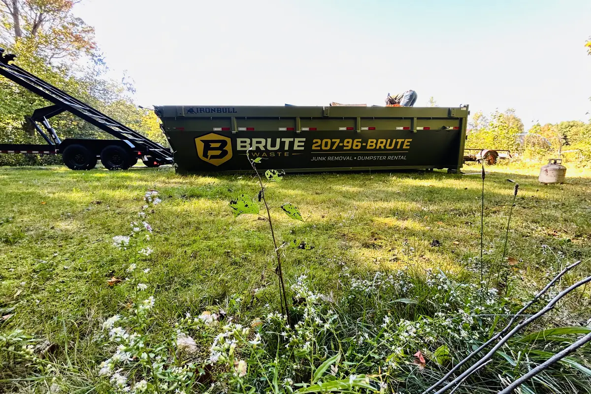 A Brute Waste roll-off dumpster standing in a rough meadow of long grass and wildflowers