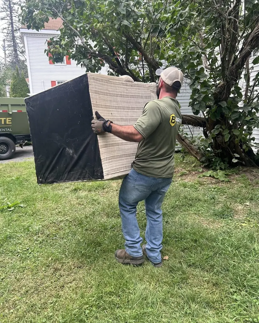 A Brute Waste crew member carrying a box spring across a lawn to the truck