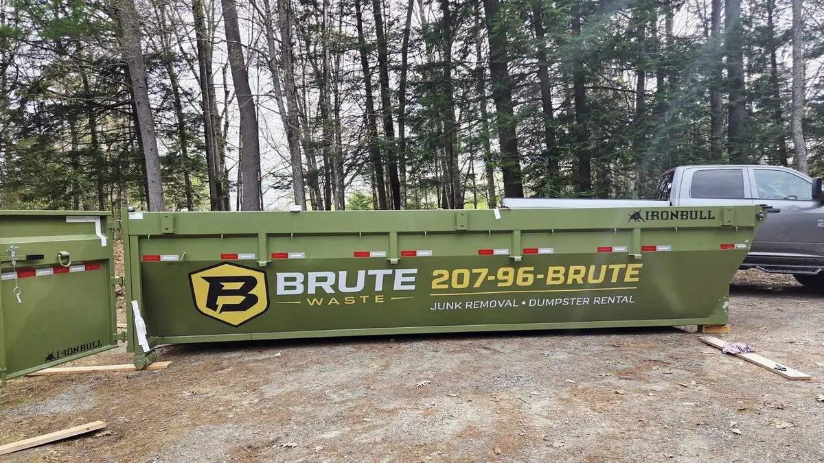 A green Brute Waste roll-off dumpster standing among tall pines in the Central Maine woods