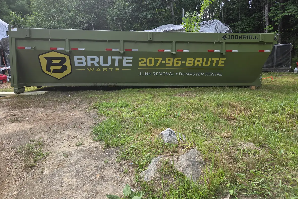 A green Brute Waste roll-off dumpster newly delivered and standing on a grass verge, the side decal clearly visible