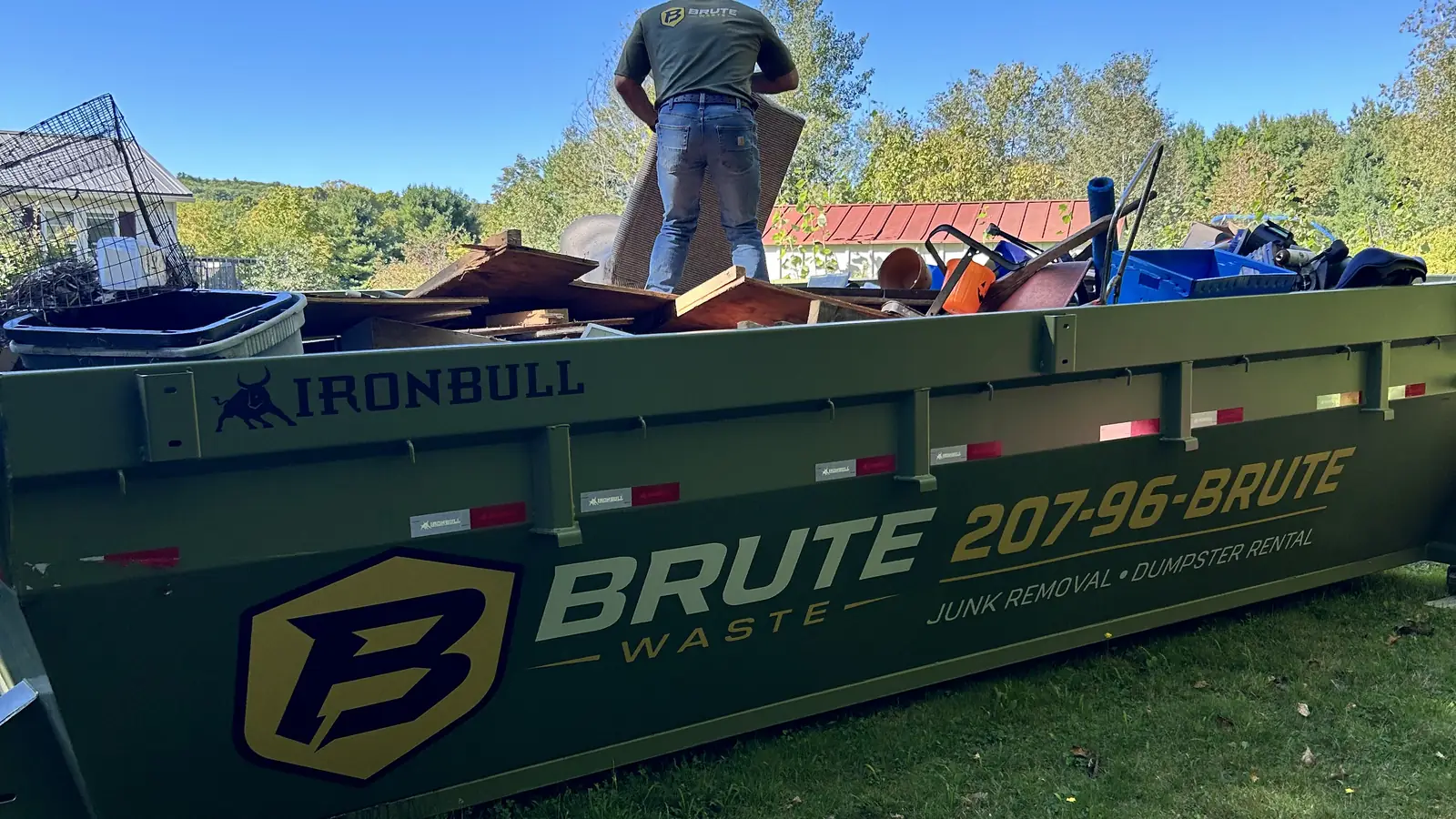 A Brute Waste crew member standing on top of a loaded roll-off dumpster against a clear blue sky