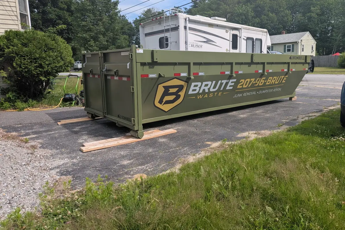 A Brute Waste roll-off dumpster set on boards on a paved driveway beside a camper