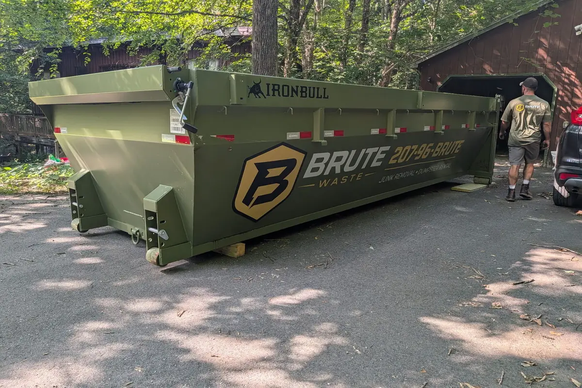 A green Brute Waste roll-off dumpster set beside a cabin on a wooded Maine lot