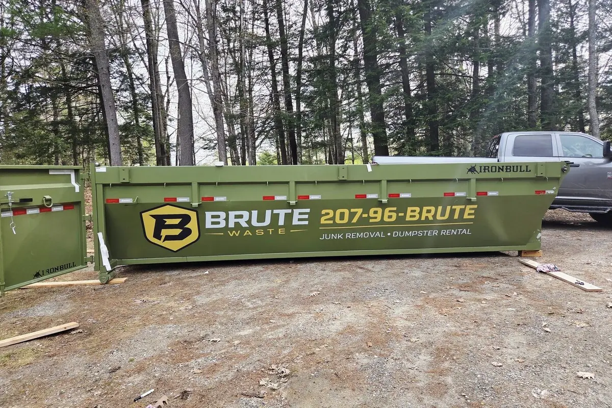 A green Brute Waste roll-off dumpster on a gravel pad among tall pines with a pickup parked behind