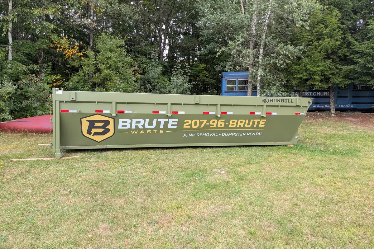 A green Brute Waste roll-off dumpster sitting on grass at the edge of a residential lot