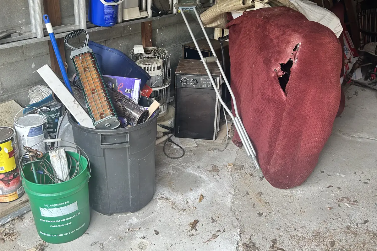 A cluttered cinder block garage packed with boxes, buckets, ladders and an upturned red canoe before a Brute Waste cleanout