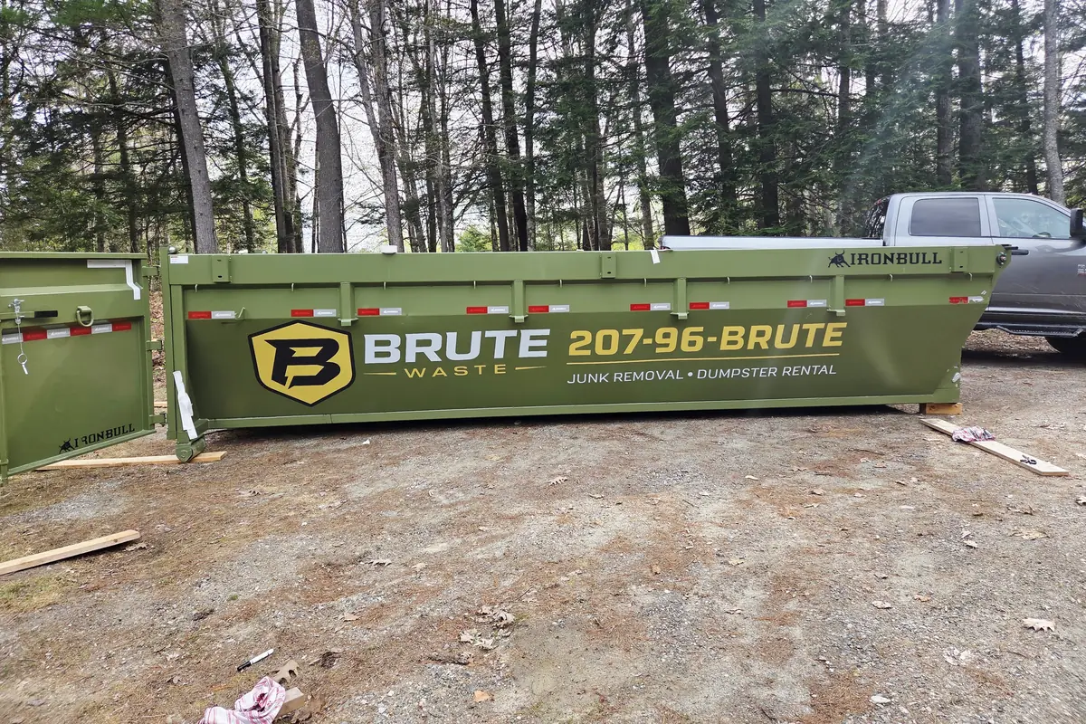 A Brute Waste roll-off dumpster set on a gravel pad with a pickup parked alongside