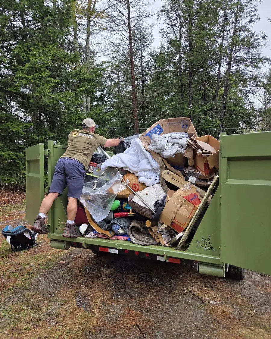 A Brute Waste crew member in a branded tee packing a household cleanout into a loaded green dumpster