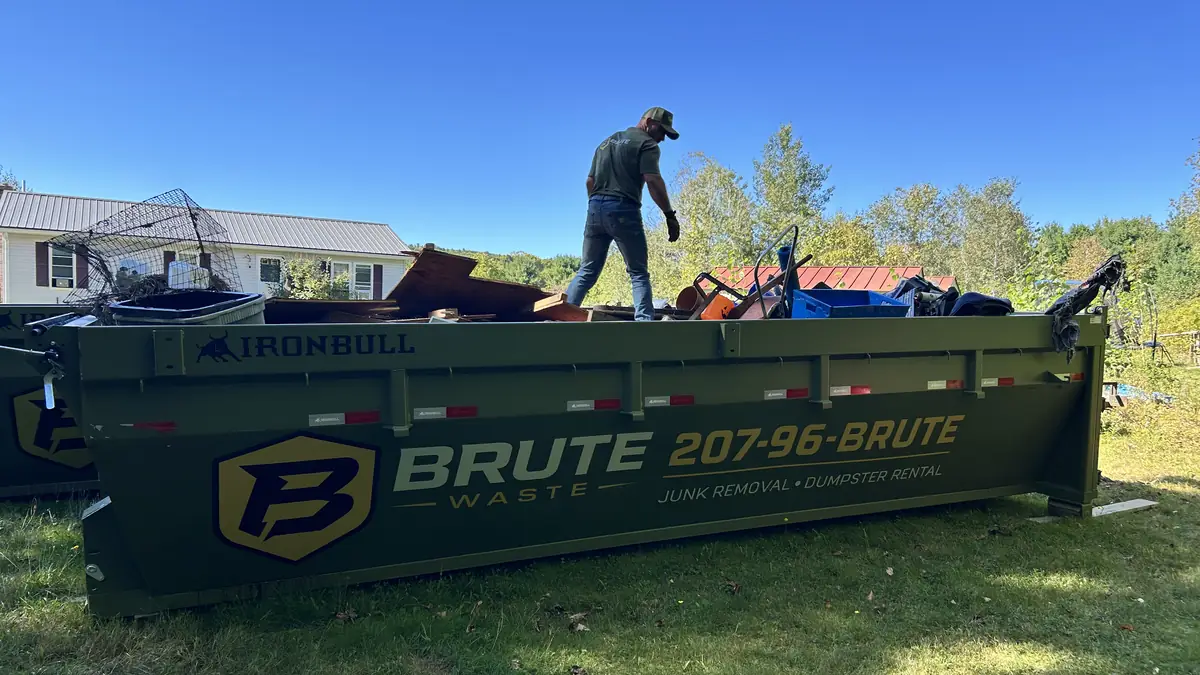A Brute Waste crew member standing on top of a loaded roll-off dumpster against a blue sky