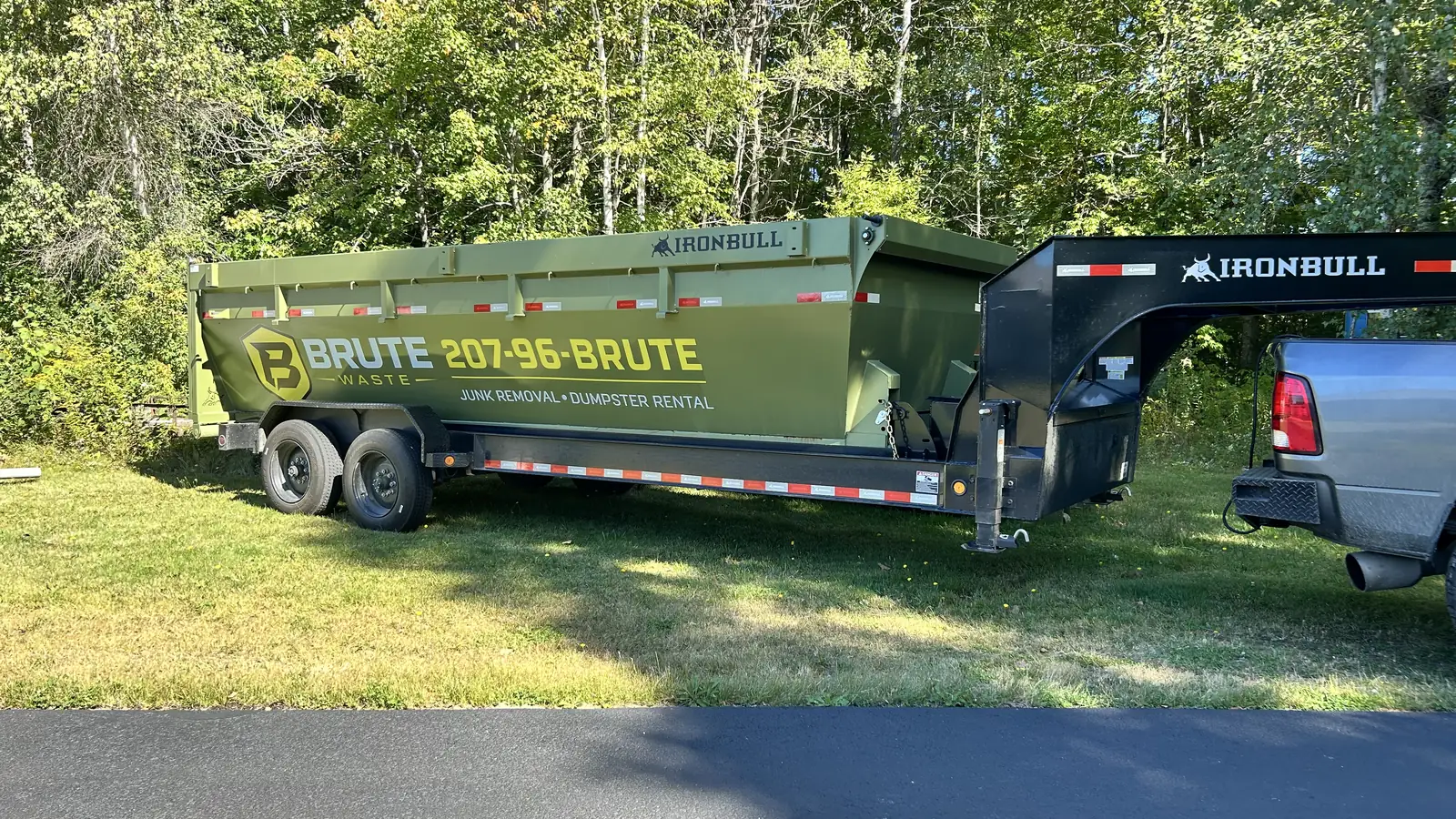 A Brute Waste dumpster and pickup parked on a sunlit residential driveway in Central Maine
