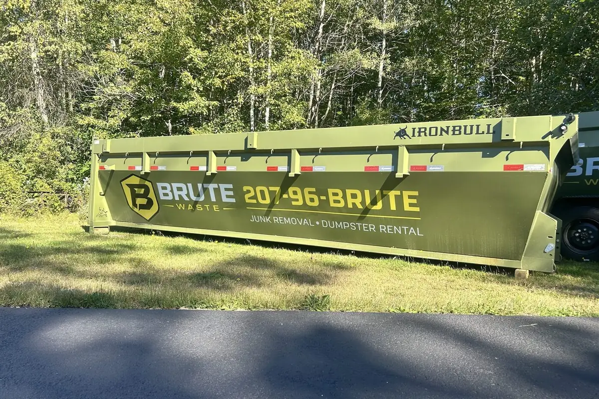 A green Brute Waste roll-off dumpster parked on grass beside a roadside treeline in Central Maine
