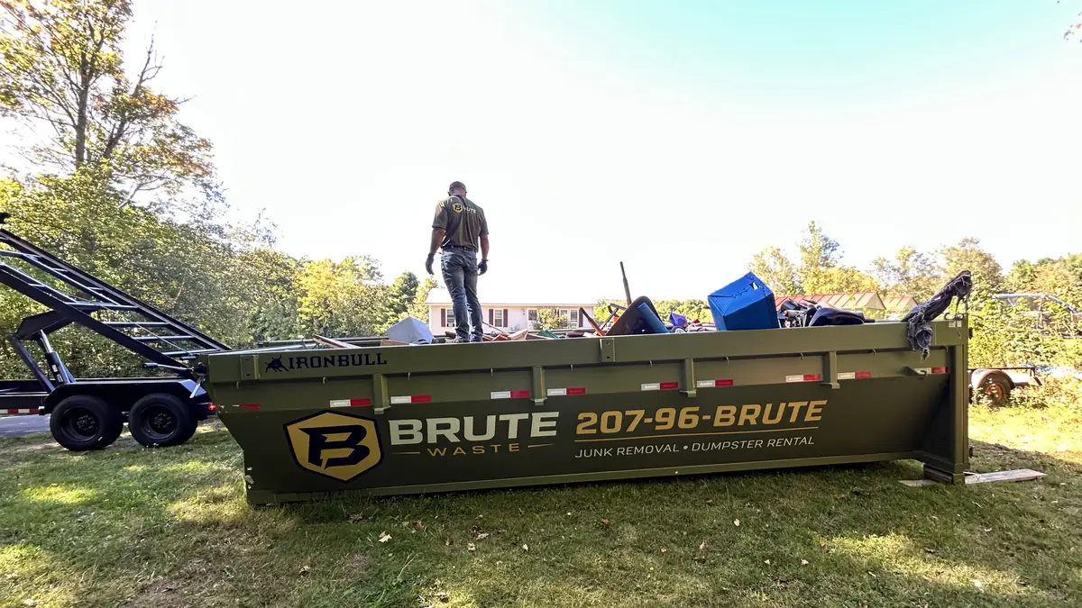 A Brute Waste crew member silhouetted on top of a loaded roll-off dumpster against a bright Maine sky