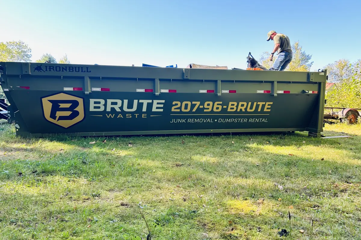 A Brute Waste crew member standing on top of a roll-off dumpster on a green lawn packing the load down