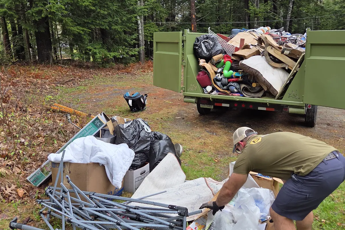 Two Brute Waste crew members lifting scrap metal frames into a loaded dumpster at a wooded job site