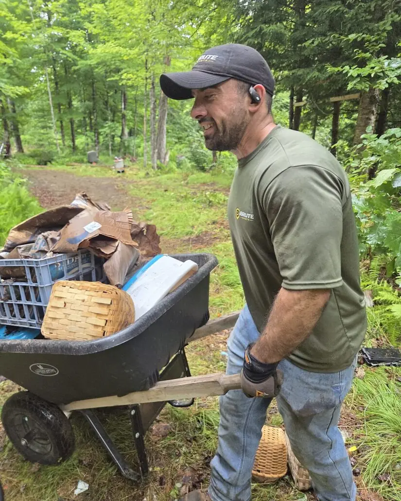 A Brute Waste crew member in a branded cap pushing a wheelbarrow of boxes along a wooded path