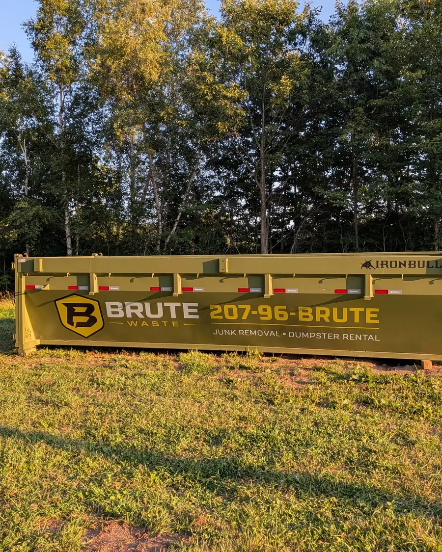 A yellow Brute Waste roll-off dumpster standing on a mown lawn with autumn trees behind it